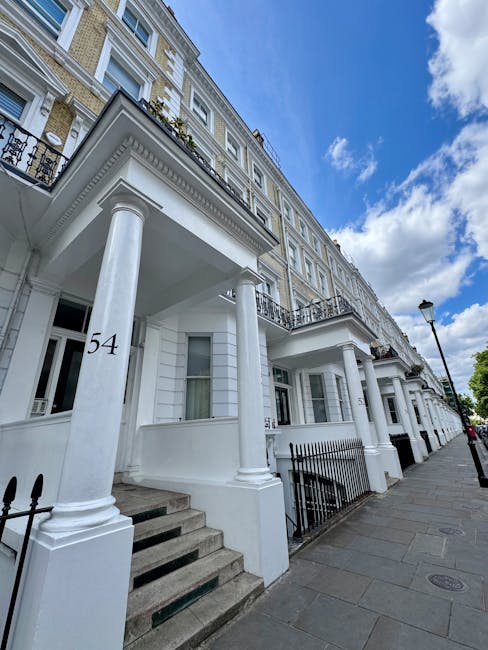 Exterior view of a white, multi-storey residential building with ornate architectural details and black wrought iron balconies. The ground level features classical columns supporting a portico, with a