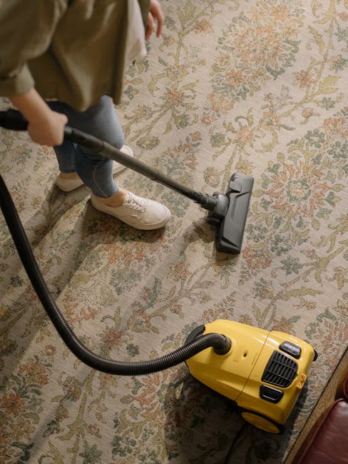 A person operating a yellow vacuum cleaner with a long black hose, cleaning a decorative patterned carpet in a residential living room. The individual is wearing beige shoes, jeans, a beige coat, and