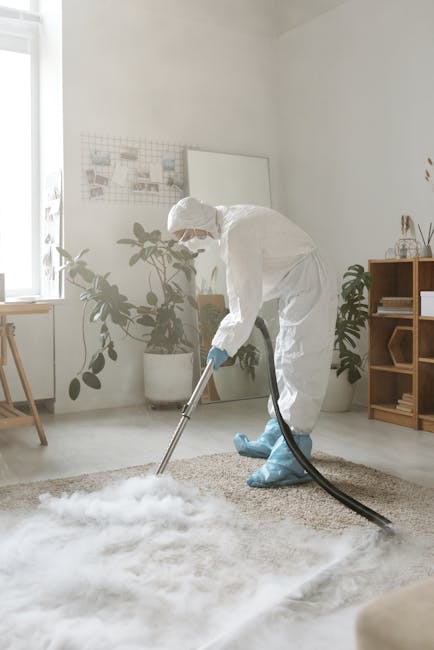 A person operating a yellow vacuum cleaner with a long black hose, cleaning a decorative patterned carpet in a residential living room. The individual is wearing beige shoes, jeans, a beige coat, and